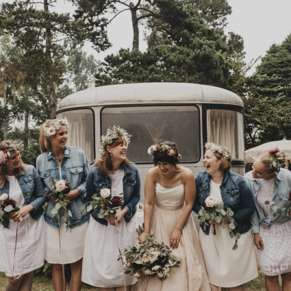 Bride laughing with bridesmaids in denim jackets and flower crowns in front of a vintage trailer